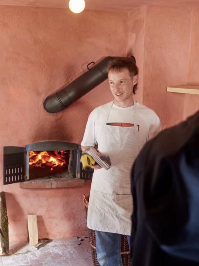 Mann in heller Schürze vor einem Ofen mit brennendem Feuer an einer rosa verputzten Wand, im Vordergrund eine Person von hinten